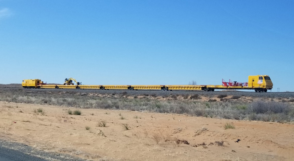 A yellow work train consisting of a squarish cab unit up front, a similar drive unit in back, and six gondolas in between, with a crane on top of the fifth gondola, is seen on a track in the desert