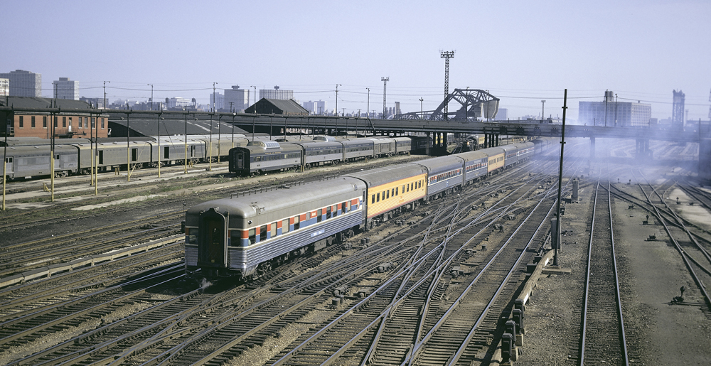 Train of stainless steel cars, except for two yellow cars, heads away from camera in yard.