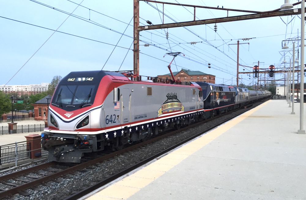 Red-and-silver electric locomotive pulling two special-paint diesels and passenger train