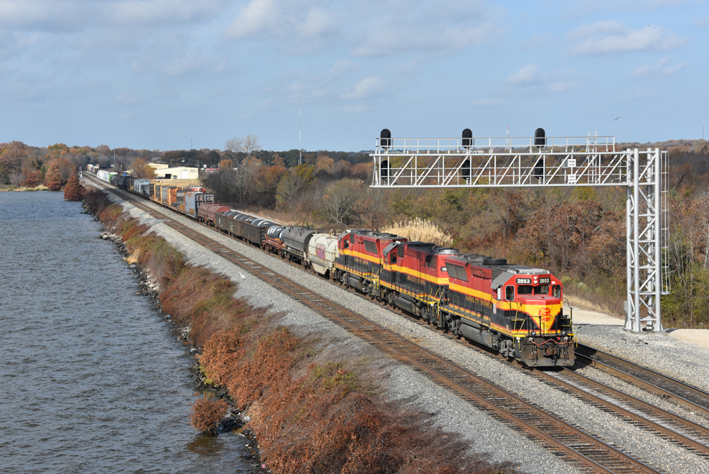 Train with three engines passes under signal bridge on track next to river.