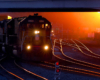 Locomotive hauls a train on yard tracks under a bridge backlit by low angle sunlight.