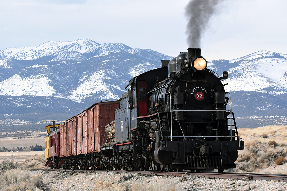 Steam locomotive hauling short freight train in an arid scene. Snowcapped mountains are in the background.