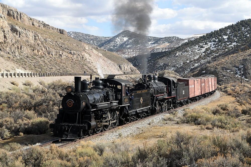 Two steam locomotive lead a freight train upgrade through a hilly valley with snow-capped mountains in the background.