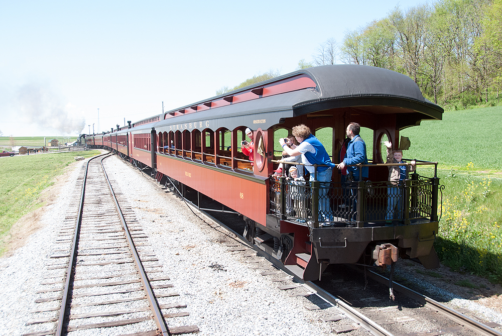 People with hand-held cameras take pictures from the rear of a moving passenger train in rolling green countryside.