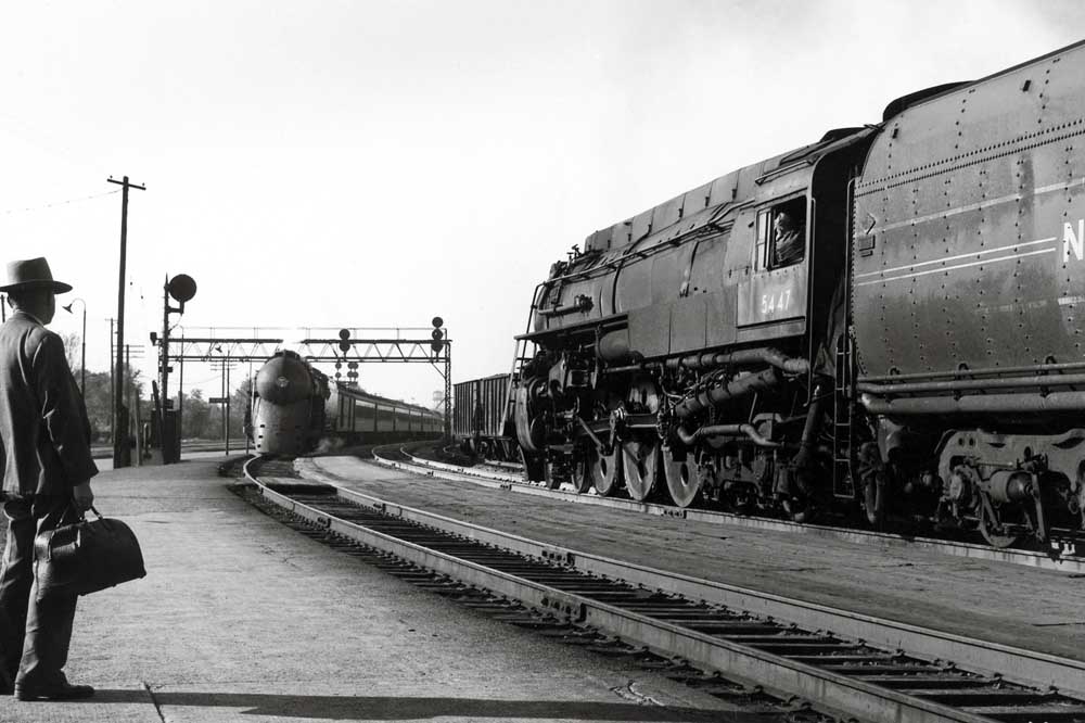 Steam-powered passenger trains meet in station on curve while a passenger watches