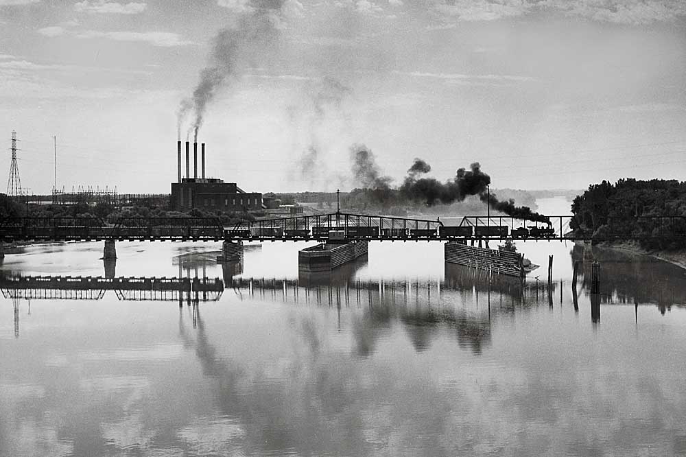 Profile of steam-powered freight train on truss bridge over wide river