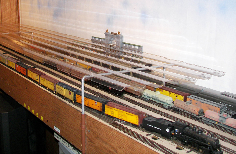 An HO scale train yard on a shelf is covered with a clear corrugated plastic dust cover