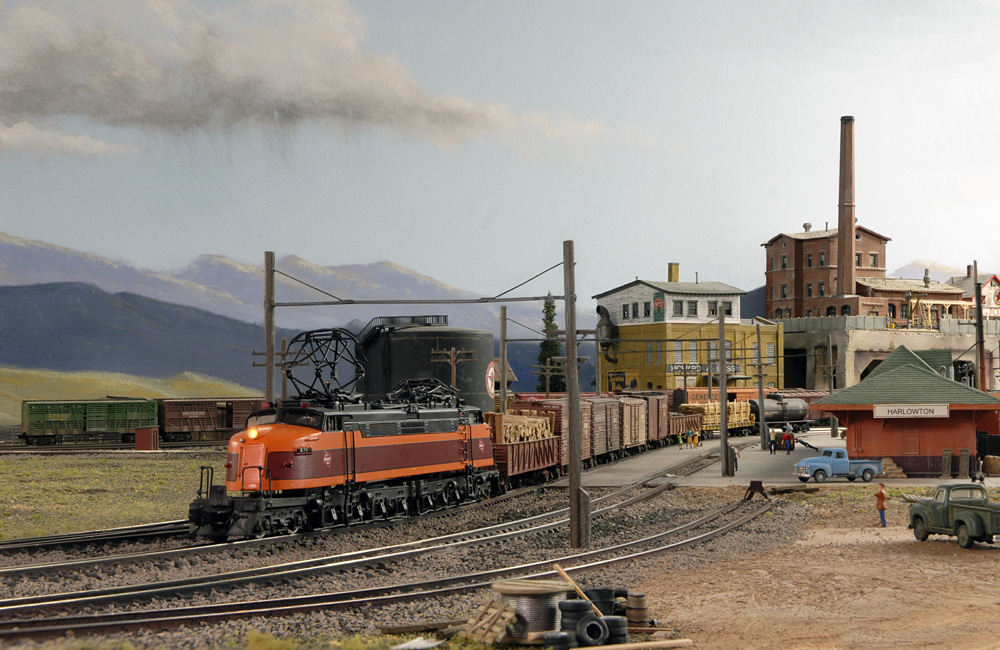 A maroon-and-orange double-ended electric locomotive couples onto a manifest train in front of a depot