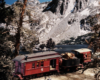 Two formally dressed figures stand beside a steam-powered one-car excursion train in a mountainous setting