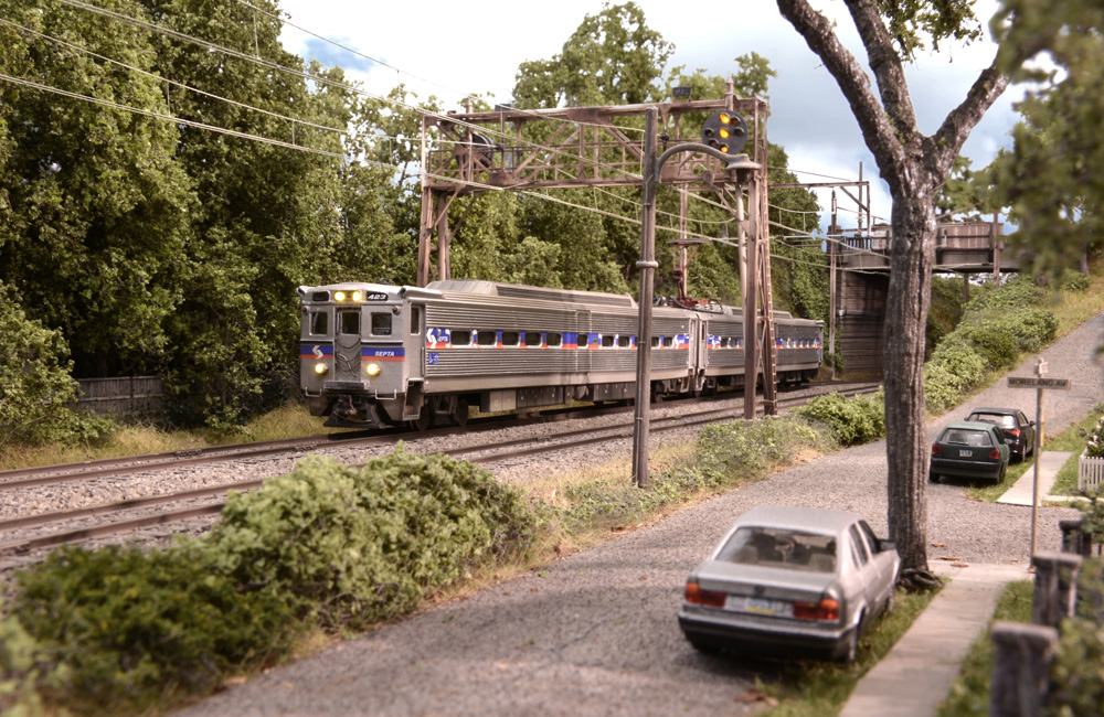 A silver two-car electric SEPTA commuter train glides under an overpass by a residential street