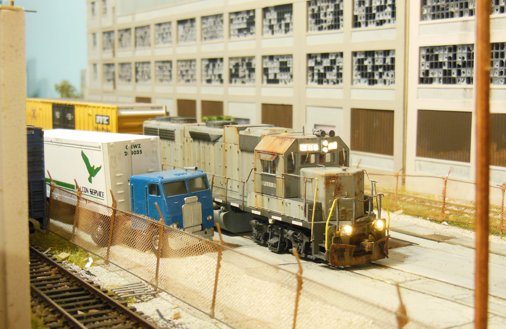 A rusty, weathered, gray diesel moves through a paved lot between tall industrial buildings