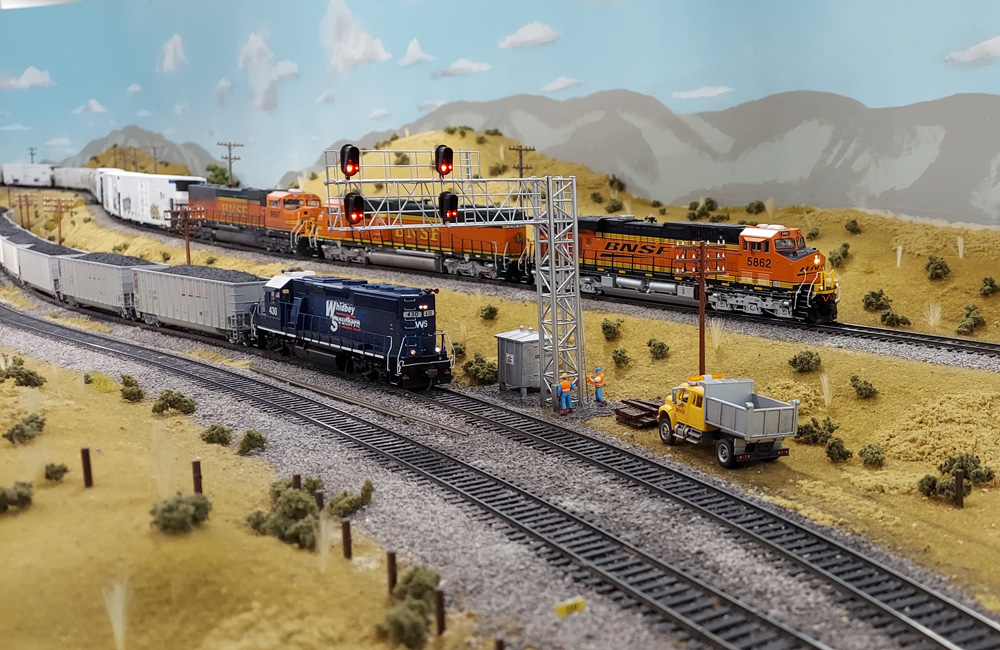 A trio of orange-and-green BNSF diesels leads a mixed freight on the high line while a blue locomotive shoves on the rear of a coal train below