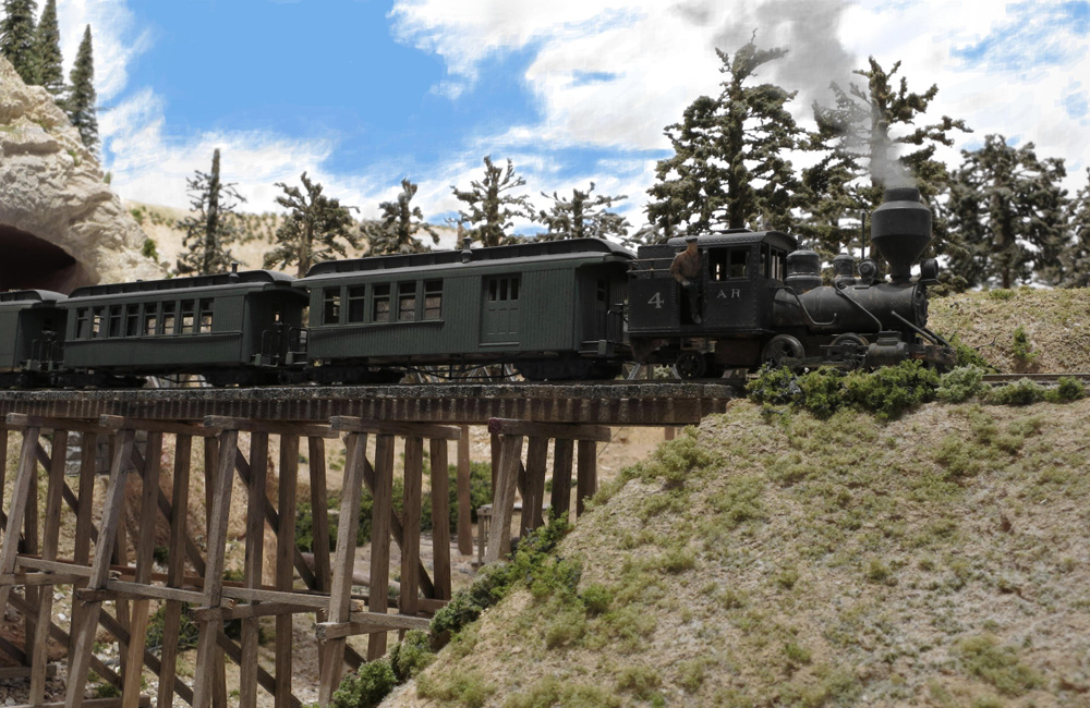 The engineer leans out the door of a small Porter steam engine as it pulls a passenger train over a wood trestle