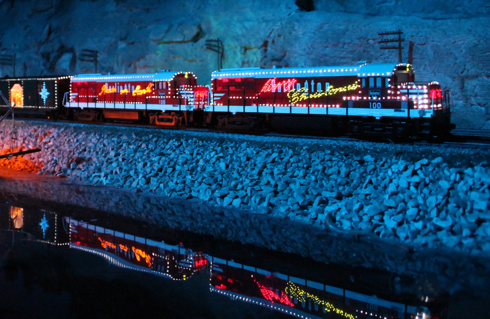 A train decorated in Christmas lights rolls along a riverbank at night, reflecting in the water