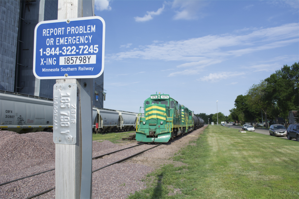 Two grade crossing identification signs on wood post with locomotives and freight cars in background.