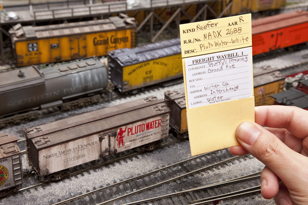 A hand holds up a car card for a refrigerator car, with a destination card in its pocket, in front of the car on an HO scale layout