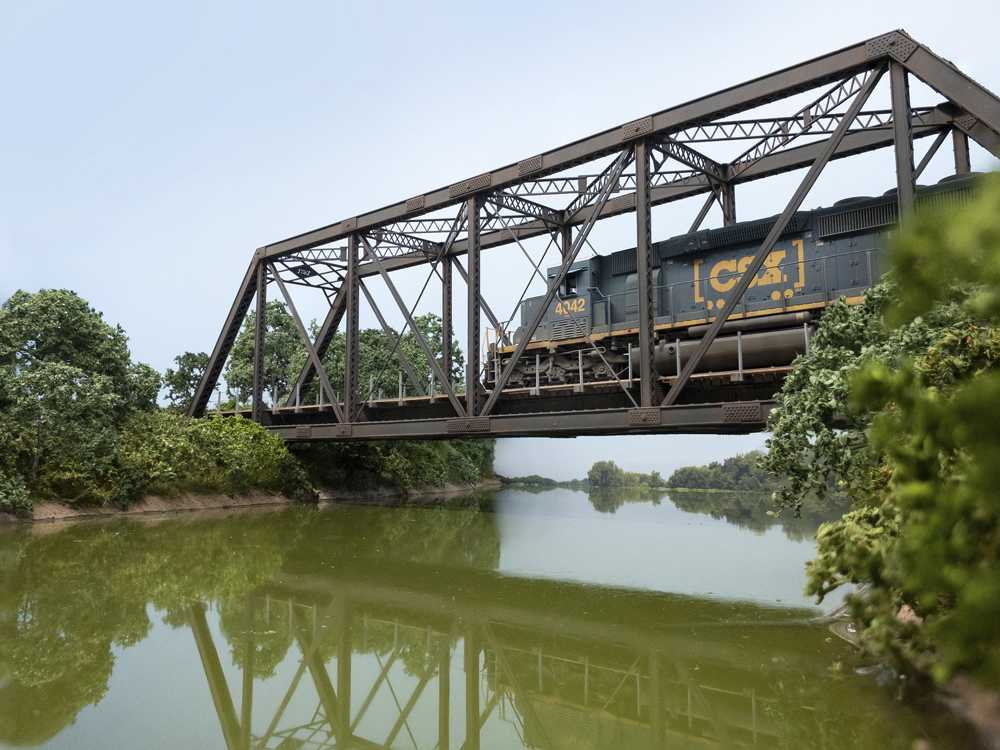 A blue CSX diesel locomotive crosses a truss bridge over a muddy green river