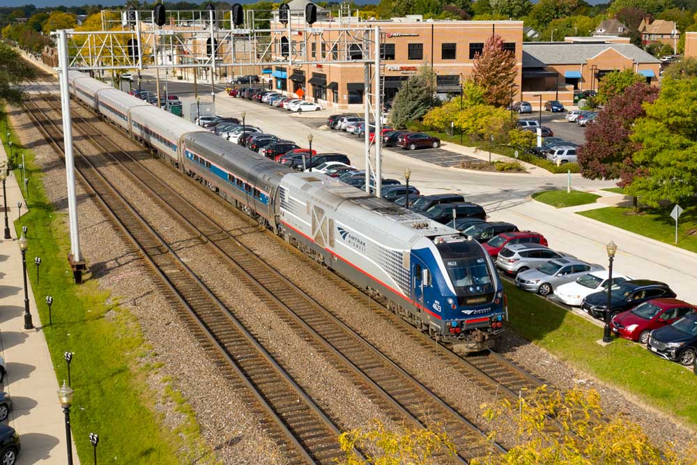 Passenger train passes under signal bridge between parked automobiles