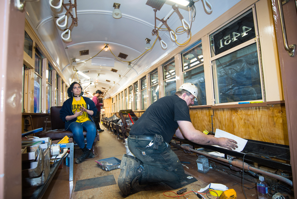 Man in hard hat, kneeling inside vintage transit car under repair .