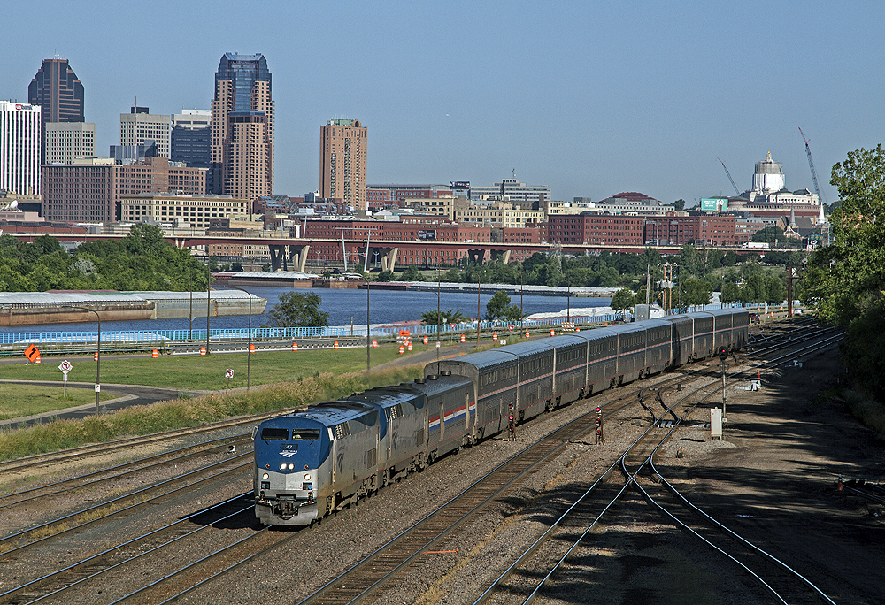 Passenger train running along flat land. A river and big-city skyline are in the middle and backgrounds.