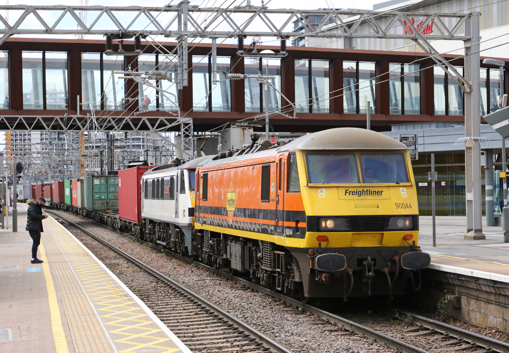 Orange and yllow locomotive leads freight train through passenger station