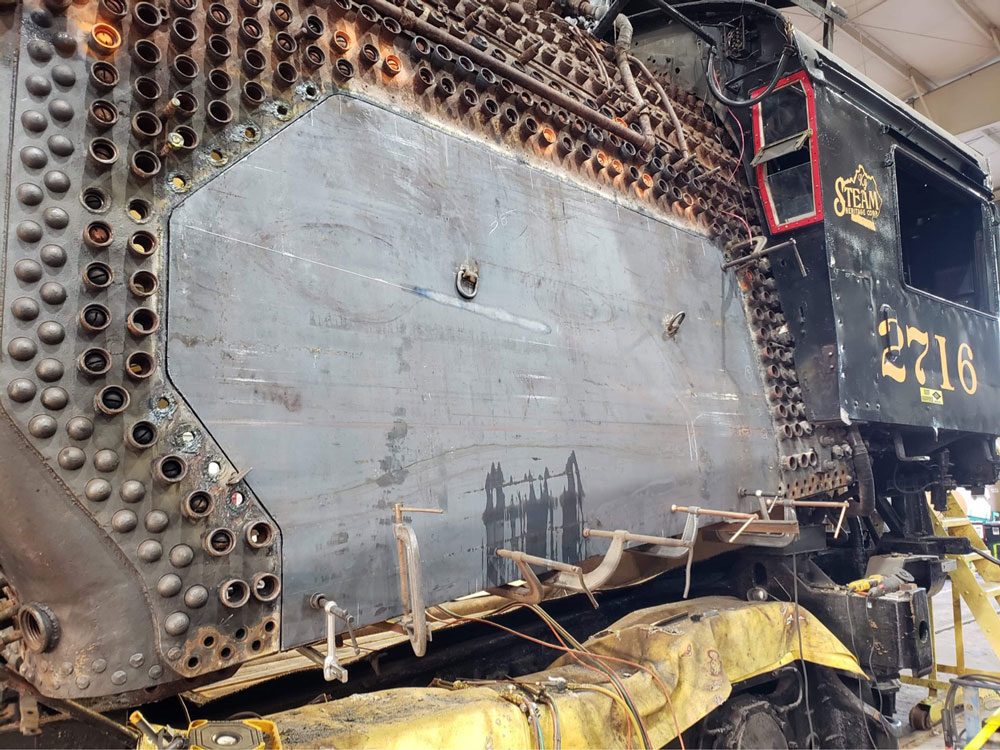 Close up of side of steam locomotive showing bare metal, plenty of holes and some riveting surrounding a large mostly flat metal piece.