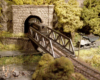 A Pennsylvania Railroad diesel switcher emerges from a tunnel onto a pony truss bridge above a road and creek