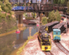 A pair of coal trains wind under a bridge and along a river toward the camera with a city scene in the background