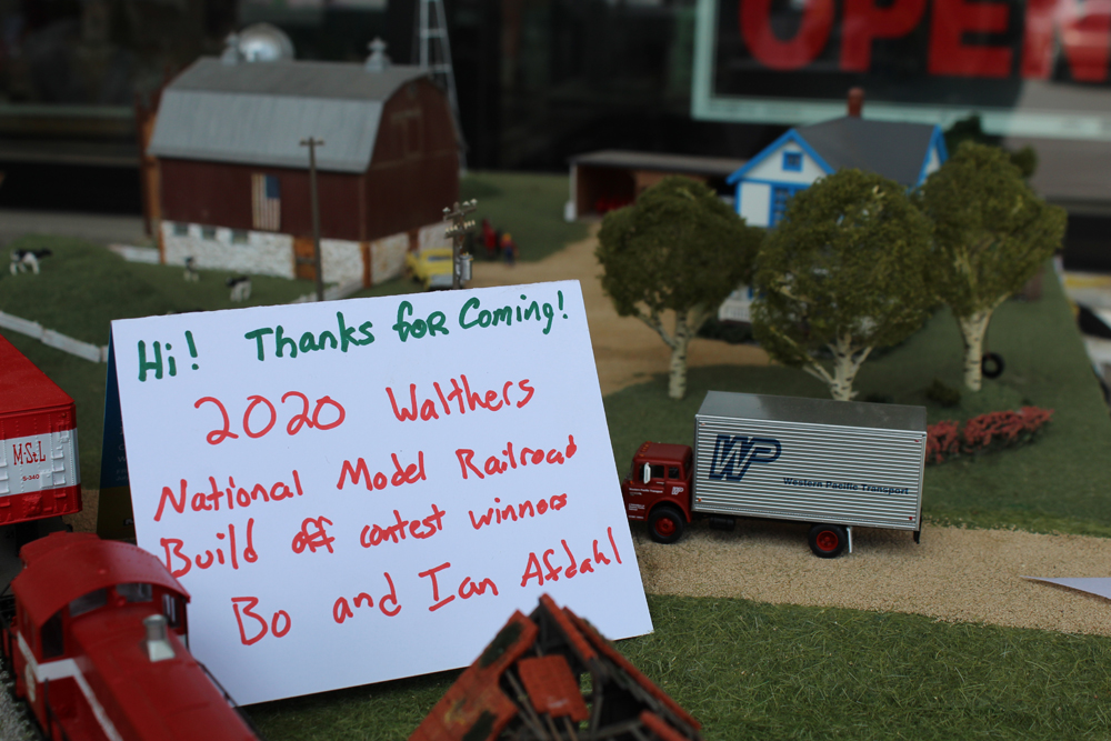 Placard resting on a model layout that says, “Hi! Thanks for coming! 2020 Walthers National Model Railroad Build off contest winners Bo and Ian Afdahl.