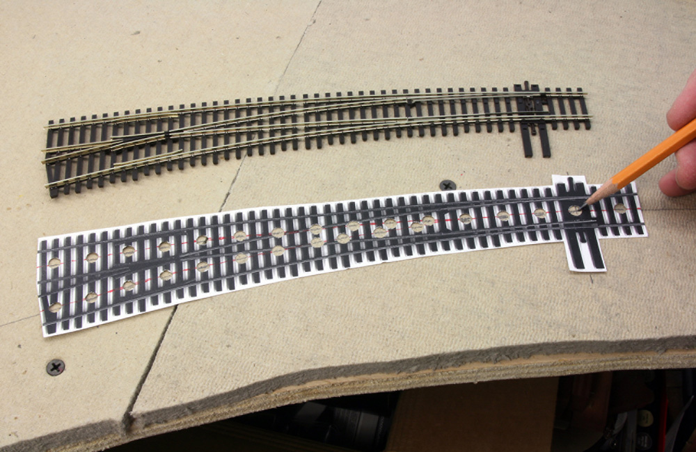 A curved HO scale turnout is seen on unfinished layout benchwork next to a paper turnout template