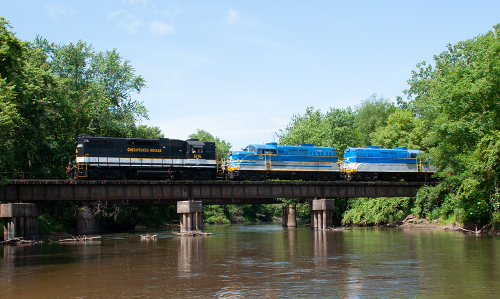 Black and white locomotive pulls two blue and white locomotives as train crosses bride