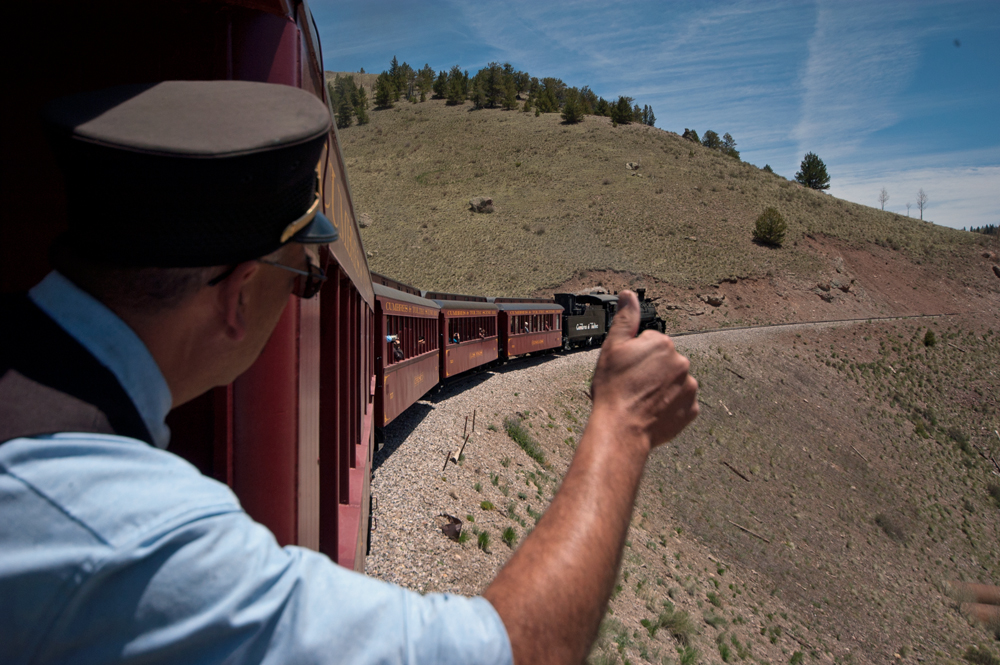 A man in rail uniform leaning over the edge of a train car to signal a locomotive crew with a Thumbs Up sign.