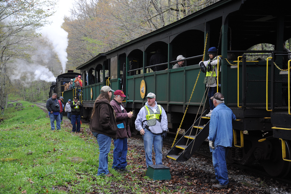 Passengers mill around near a stopped tourist train with open-air cars.