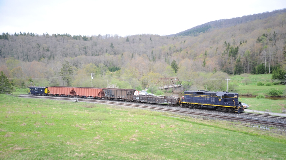 Freight train with blue and yellow diesel, four cars, and a caboose, rolls through a field by a river with mountains as a backdrop.