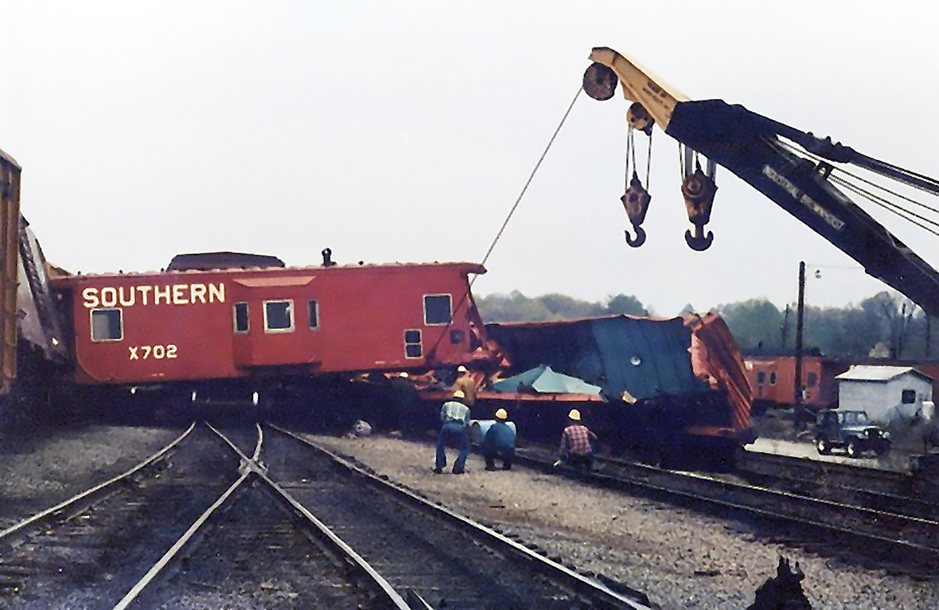 A red caboose sitting perpendicular over railroad track tied to a crane on an overcast day.