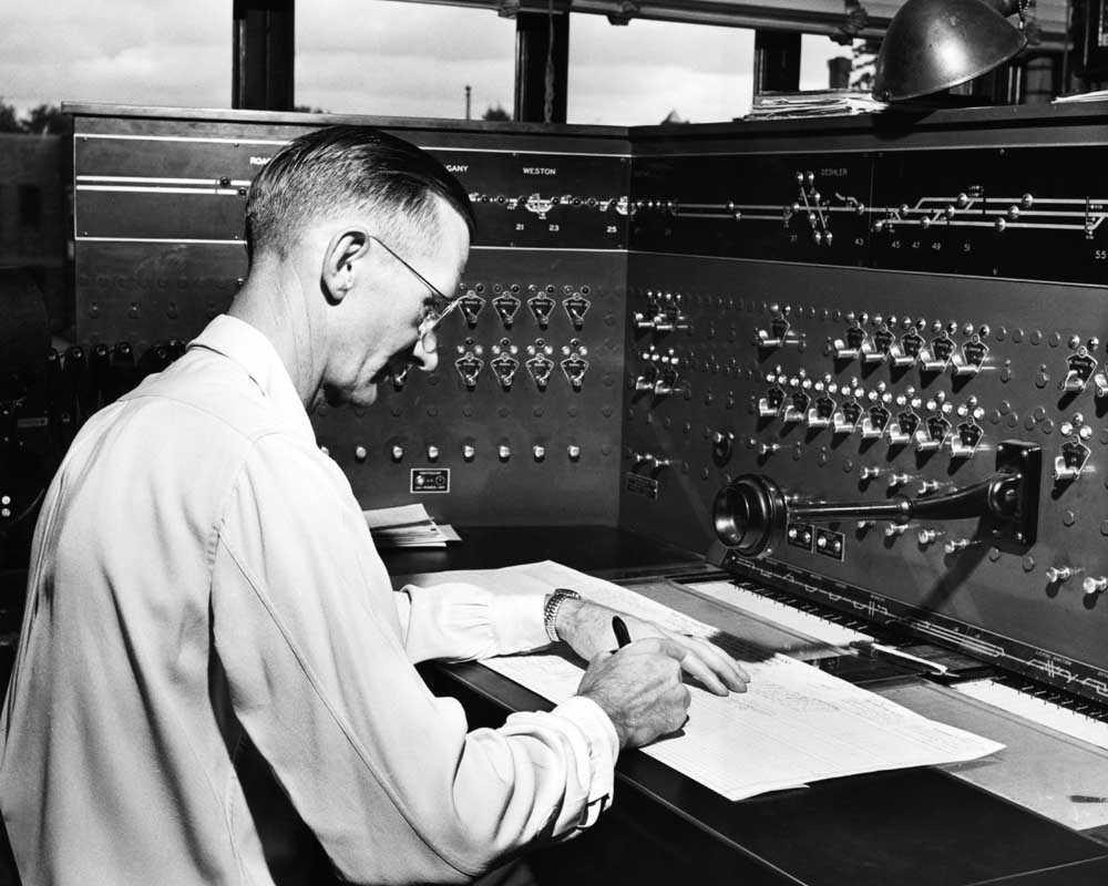 Man sits in front of dispatchers’ panel making notation on paper