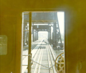 Bridge structures and a double-track appear outside of a caboose door.