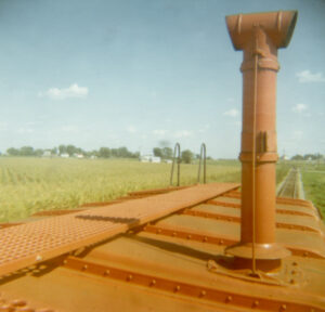 Orange-red smokestack, roof walk, and a ladder end appear in the foreground against a backdrop of row crops and blue sky filled with distant fluffy clouds. 