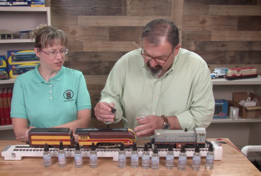 Two people with toy trains apply smoke fluid to a model steam locomotive.