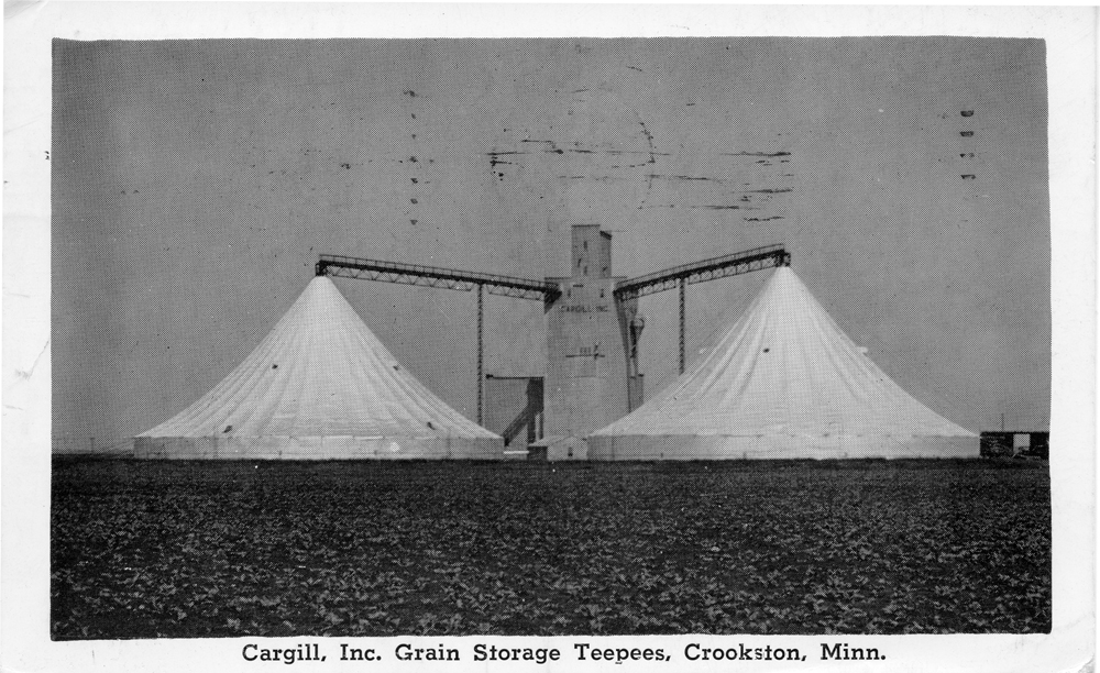 Black-and-white image of metal grain elevator with two grain storage tents and farm field in foreground.