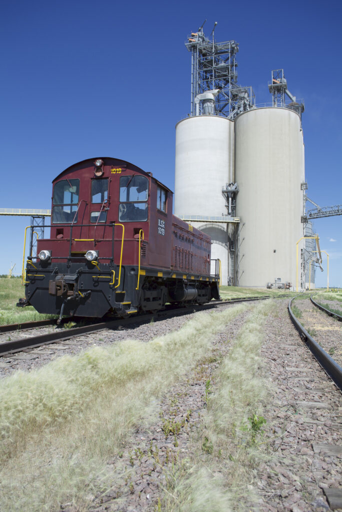 An end-cab switcher painted maroon and black on a siding next to a grain elevator complex.