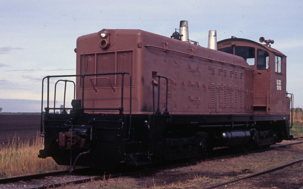 An end-cab switcher in brown primer and black paint sits on a track next to a farm field.