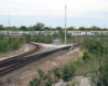 Four-car stainless steel electrified commuter train, partially obscured by brush, stops at station.