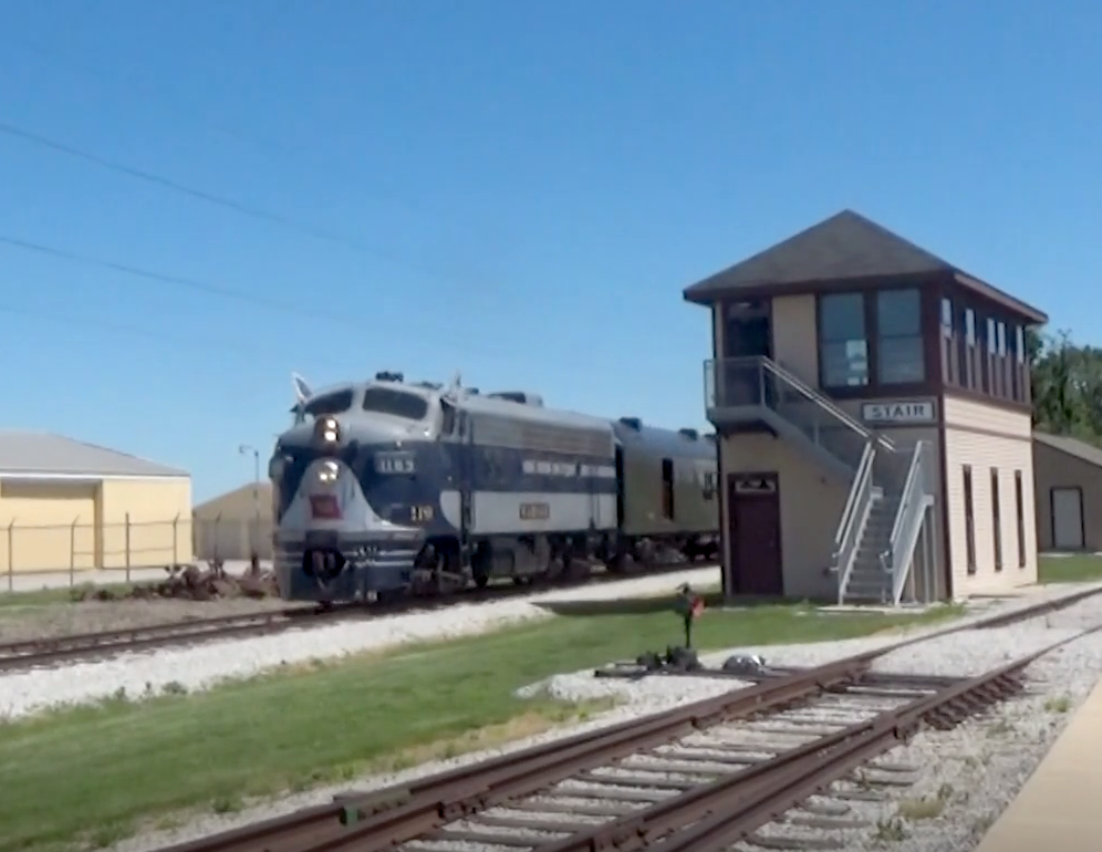A two-tone blue locomotive pulls a freight train past a railroad tower on a clear, sunny day.