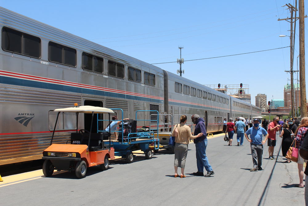 Passenger cars at station with baggage being unloaded onto trailer and passengers walking on platform