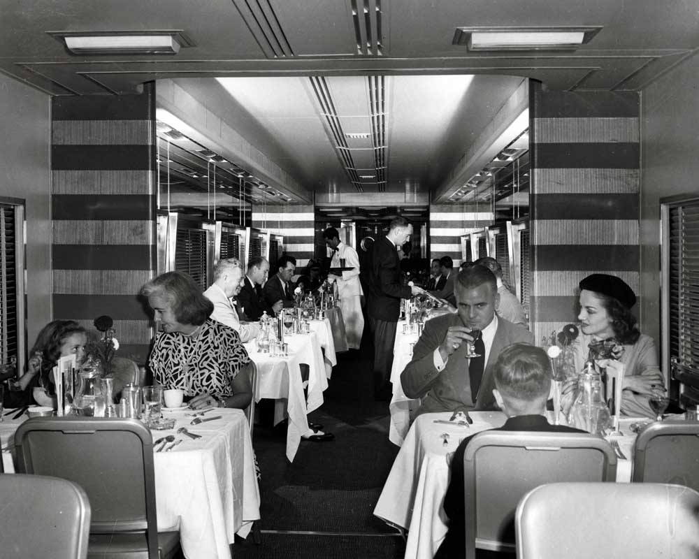 People seated at tables with cloth tablecloths on railroad dining car. New York Central passenger trains.
