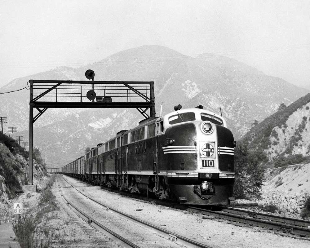 Diesel locomotive set leads a freight train through hills and under a signal bridge