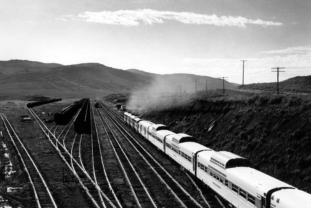 Streamlined passenger train rolls into valley alongside yard tracks filled with hoppers