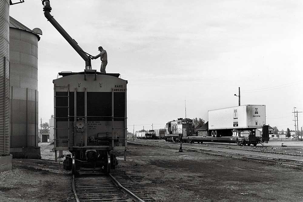 A man guides loading chute into covered hopper while short freight train passes