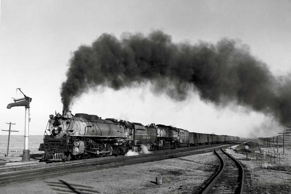 Smoking steam locomotive leads a passenger train across open terrain by a water spout